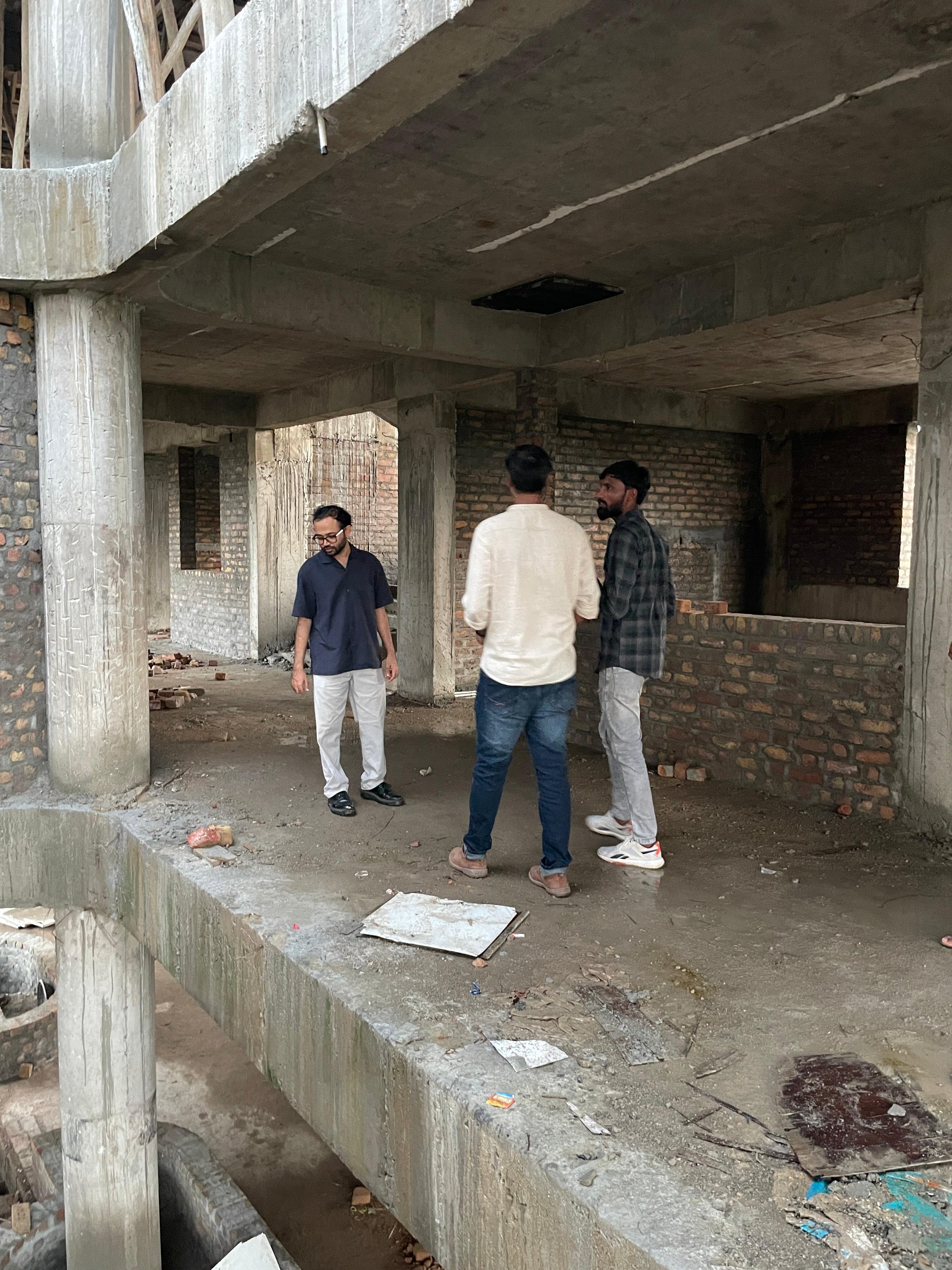 Three men standing on a concrete floor within an unfinished building with brick walls and exposed columns.
