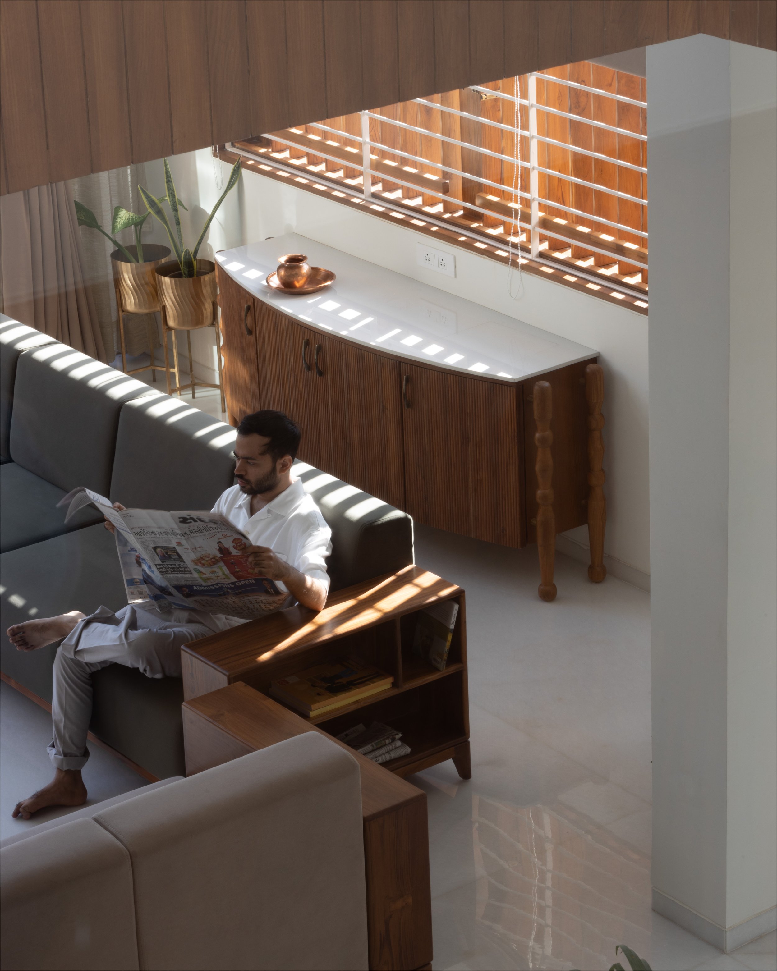Man sitting on a sofa reading a newspaper in a modern living room with wooden furniture and potted plants.
