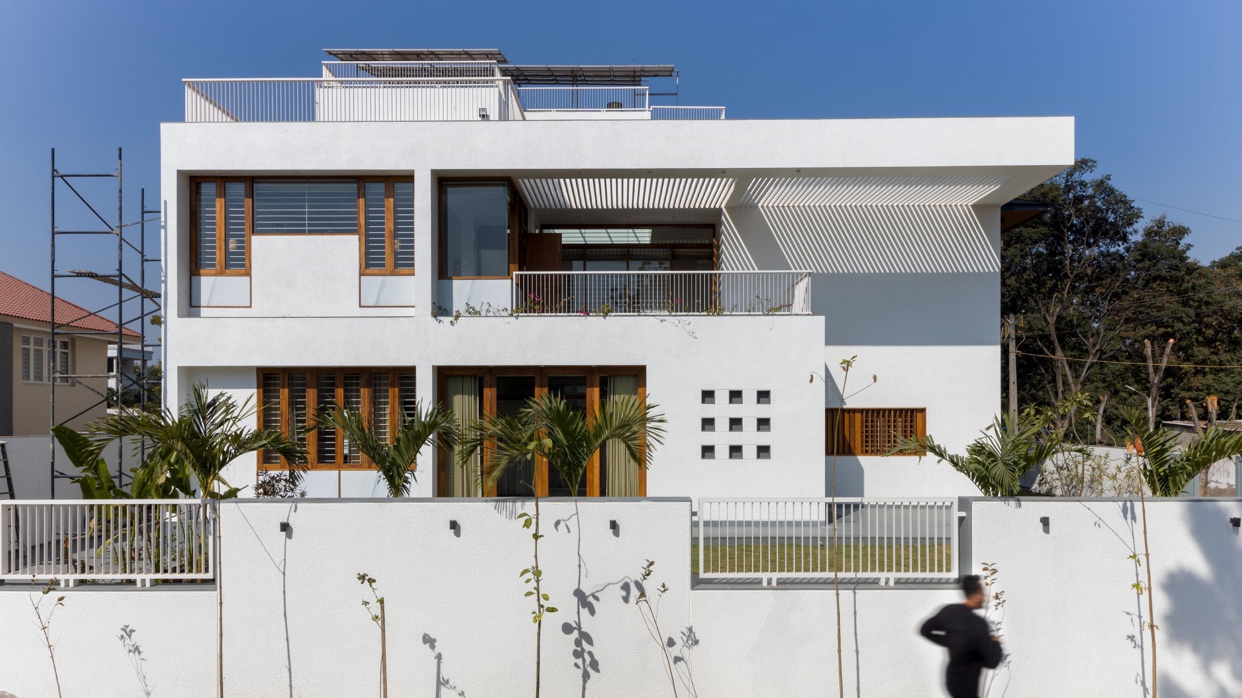 Modern white house with large windows and wooden accents, featuring a balcony with plants, surrounded by a white fence and palm trees. A person is jogging in front of the house.