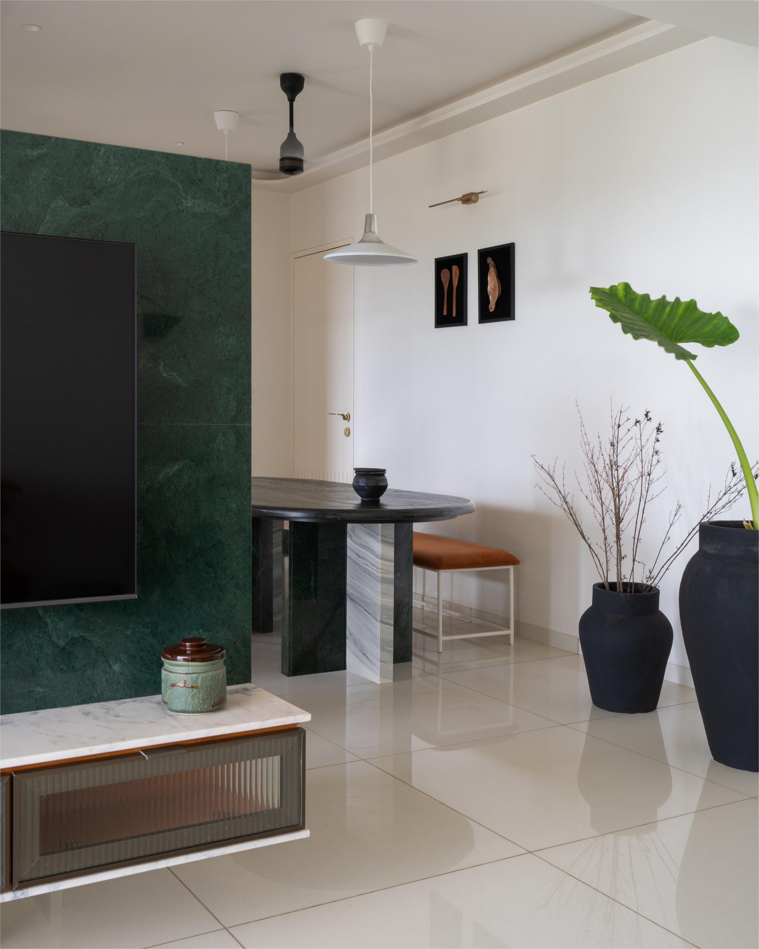 Modern living room with a green accent wall, large TV, and marble table. Minimalist decor includes black planters, a wooden bench, and framed art. Features pendant lighting and sleek, glossy flooring.