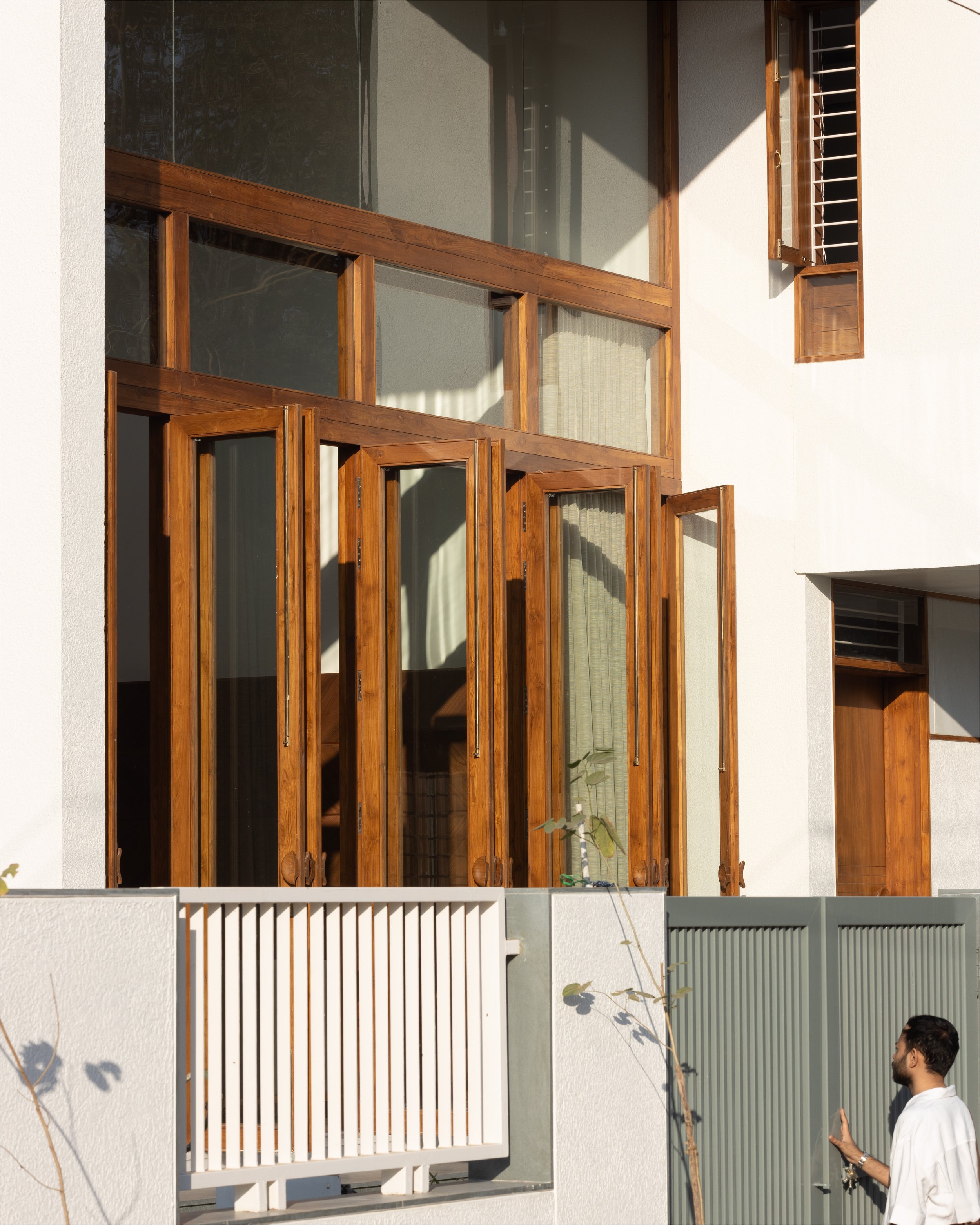 Modern house exterior with large wooden-framed windows, a white railing, and a young man standing in front of a green gate.