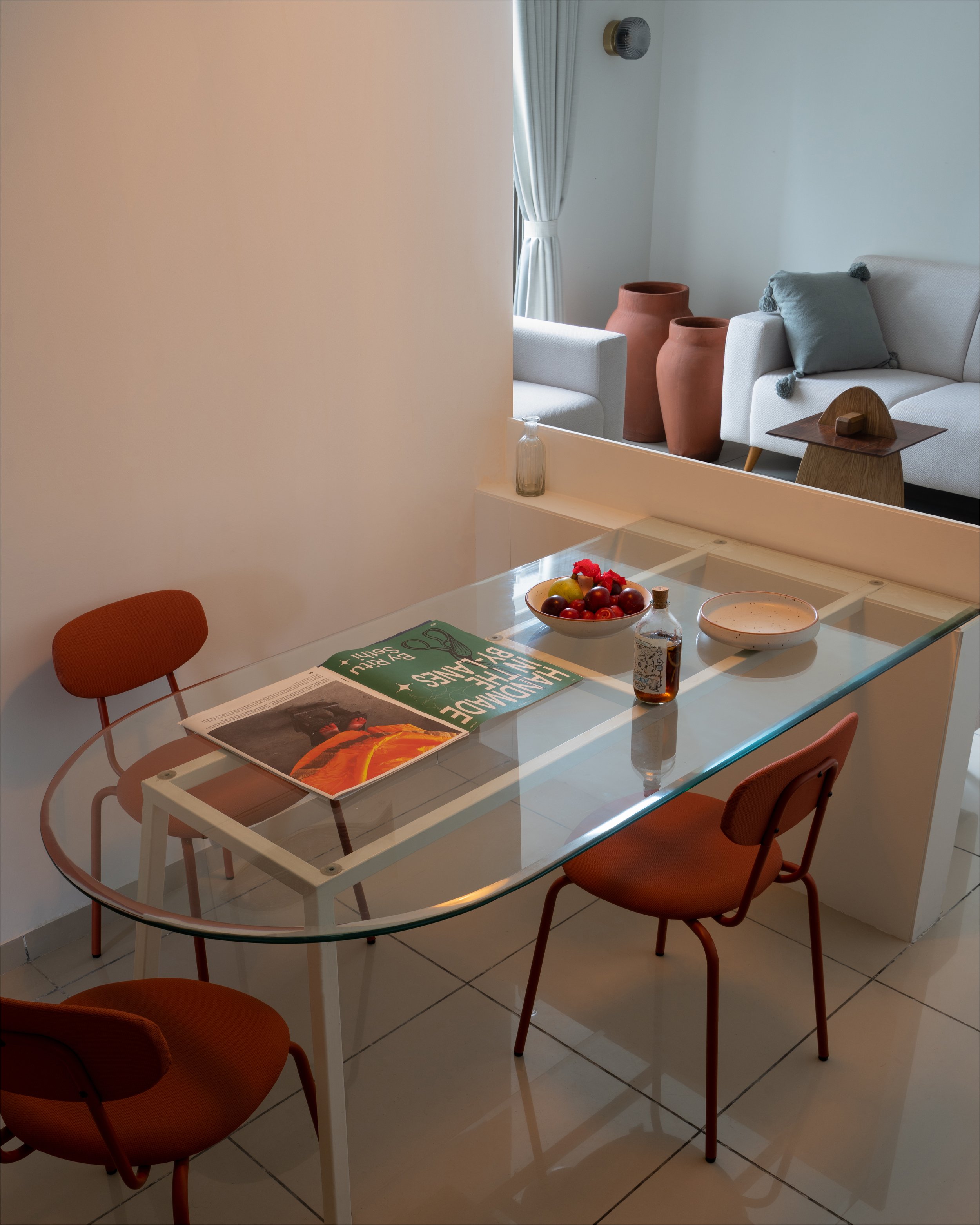 Glass dining table with orange chairs, a magazine, bowl of fruit, and a small bottle. A sofa with cushions and terracotta vases are visible in the background.