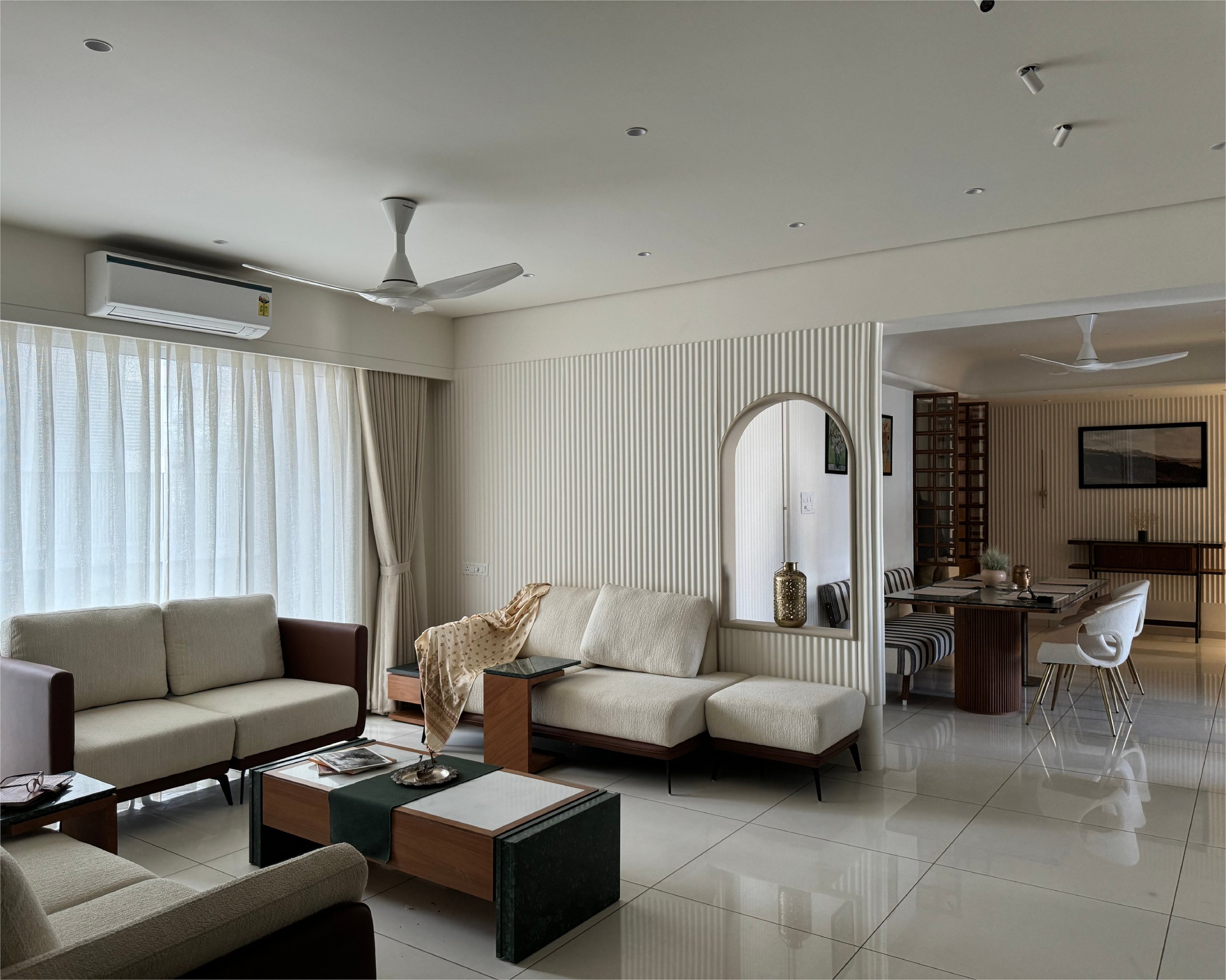 Modern living room with beige sofas, coffee table, and arc-shaped wall opening; visible dining area in background with table and chairs. Minimalist decor, tiled flooring, air conditioning unit, ceiling fans, and curtains.