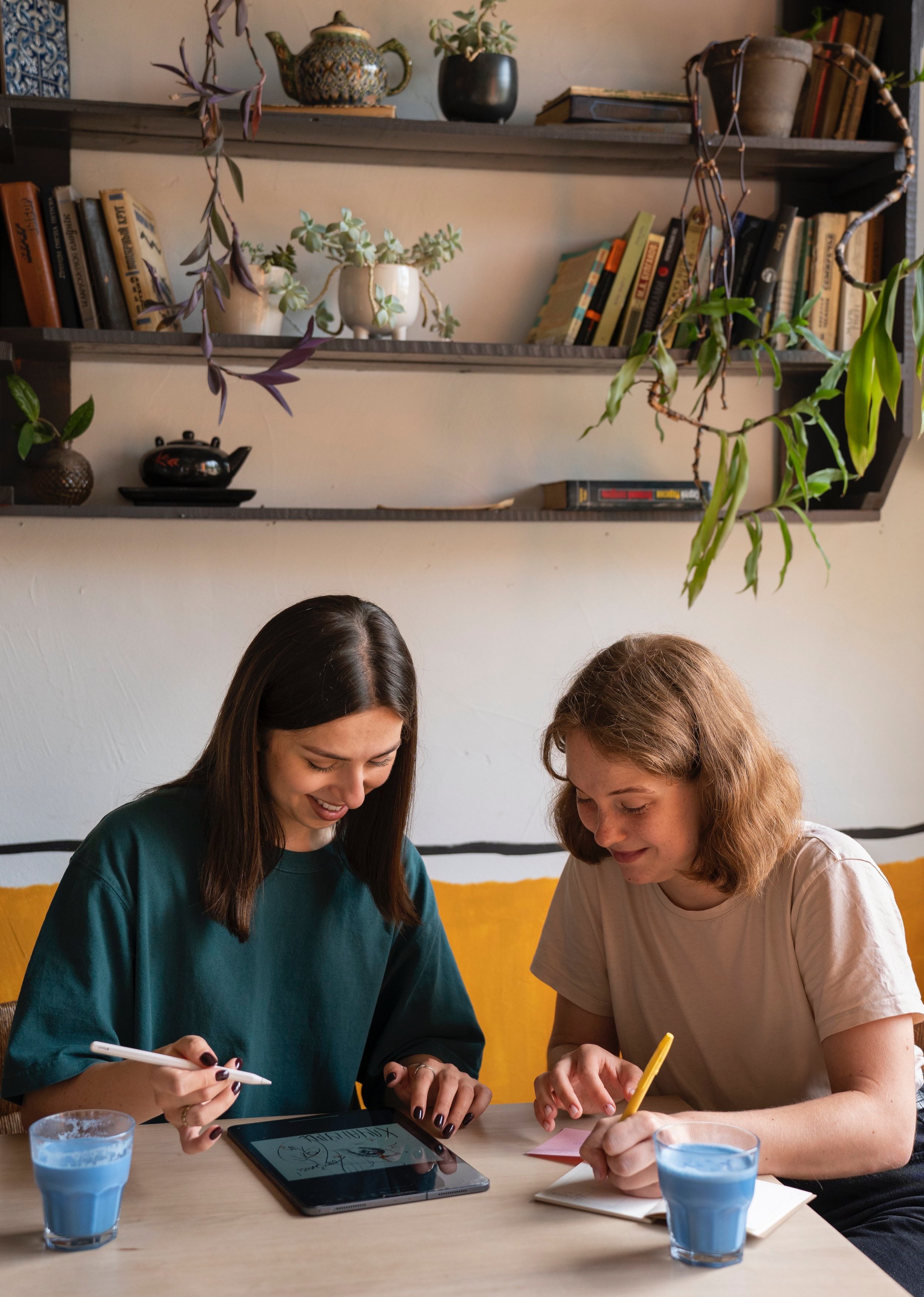 Two women sitting at a table in a cozy setting, using a tablet and writing in a notebook. Shelves with books and plants are in the background, and they each have a glass of blue drink on the table.