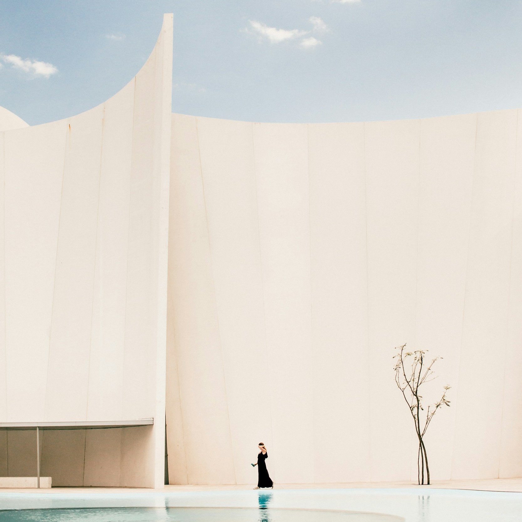 Person in black dress walking near a curved, modern white building with minimalistic architecture and a small tree.