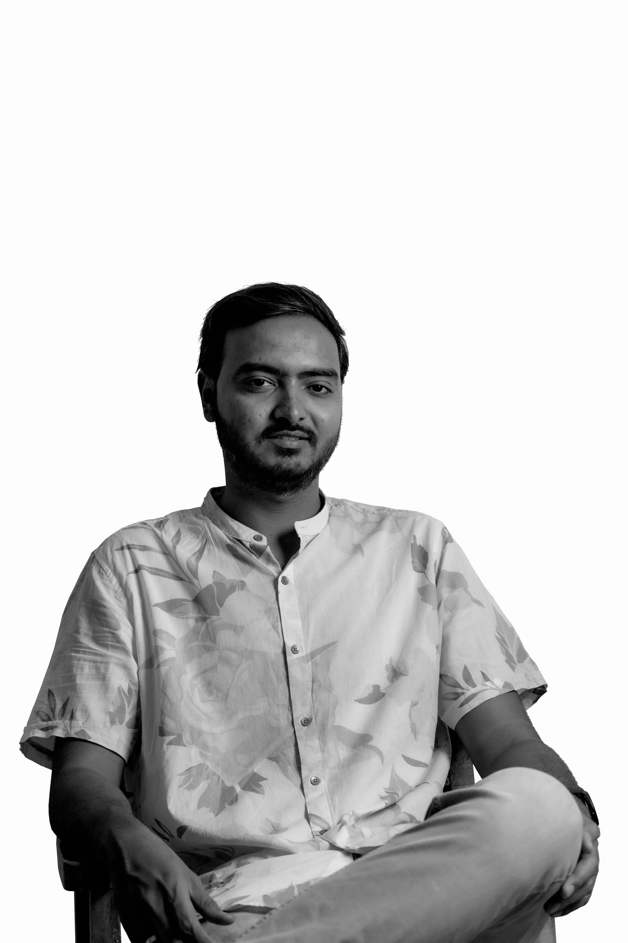 Black and white portrait of a man sitting in a chair, wearing a floral shirt, with a neutral expression against a plain background.