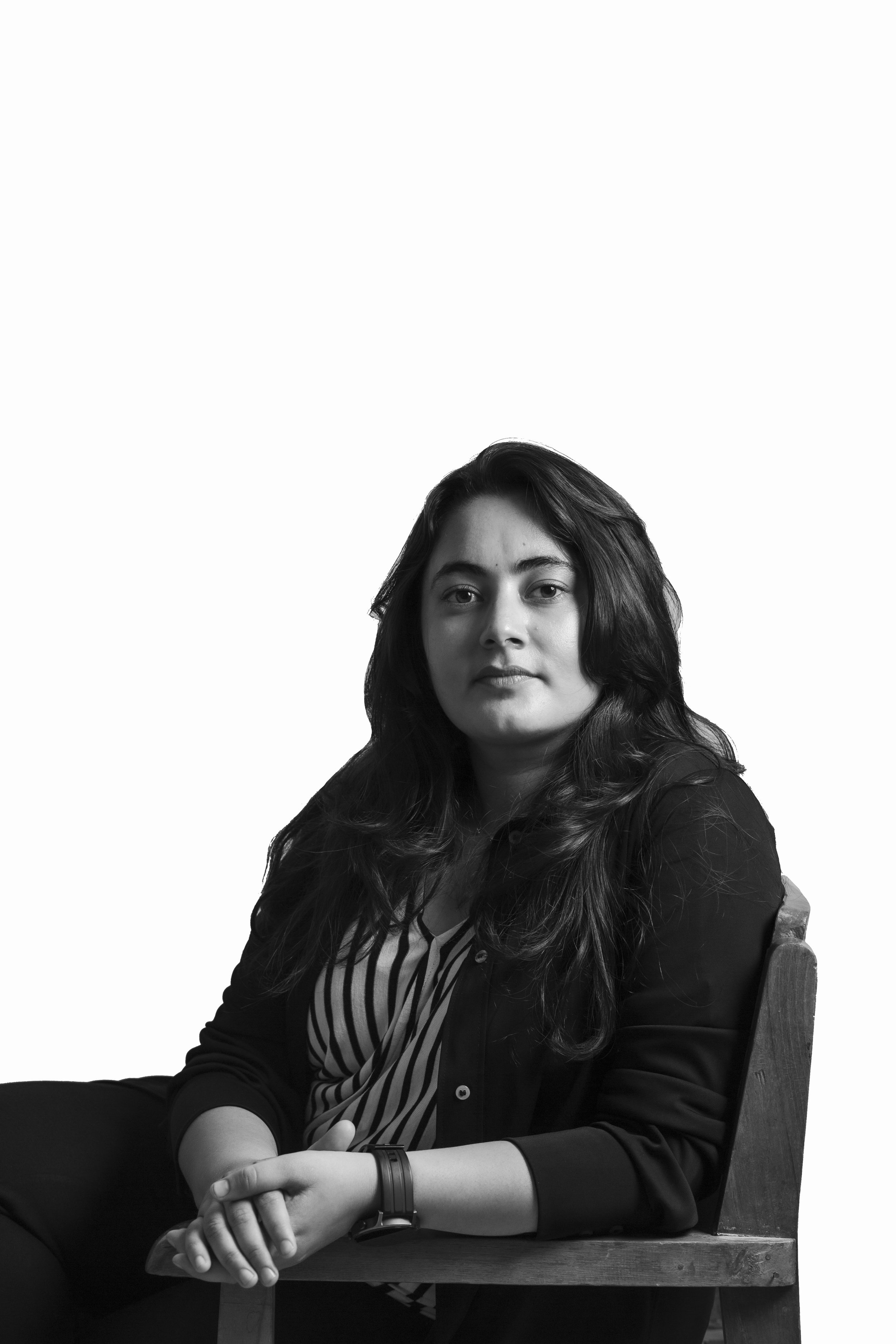 Black and white portrait of a woman with long hair seated on a chair, wearing a striped shirt and a smartwatch, looking at the camera.